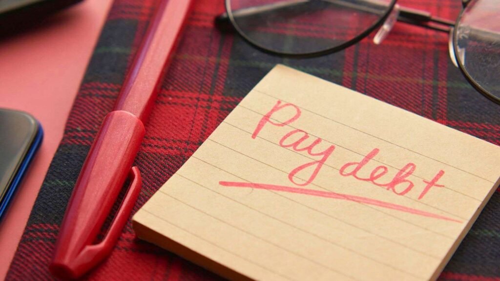 A handwritten note reading "Pay debt" sits on a desk with a red pen and glasses, illustrating the personal impact of the U.S. national debt surpassing $39 trillion.